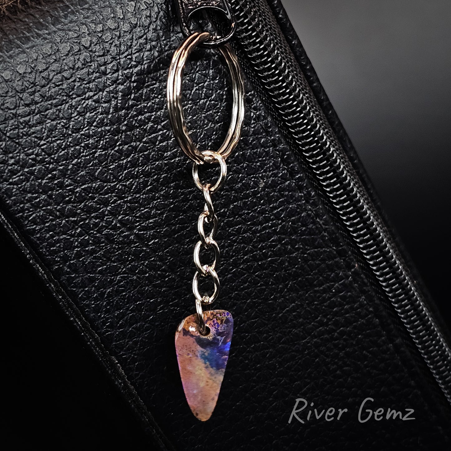 A close-up of a silver metal bag charm with a dangling iridescent boulder opal shaped like a guitar pick, resting on a black textured leather surface. The brand name 'River Gemz' is visible in the bottom right corner.