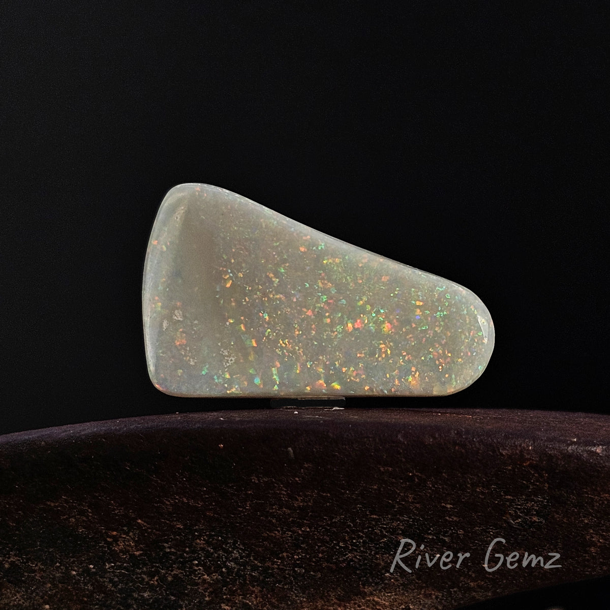 White opal on a brown surface and black background together with 'River Gemz' water mark in the bottom right hand corner.