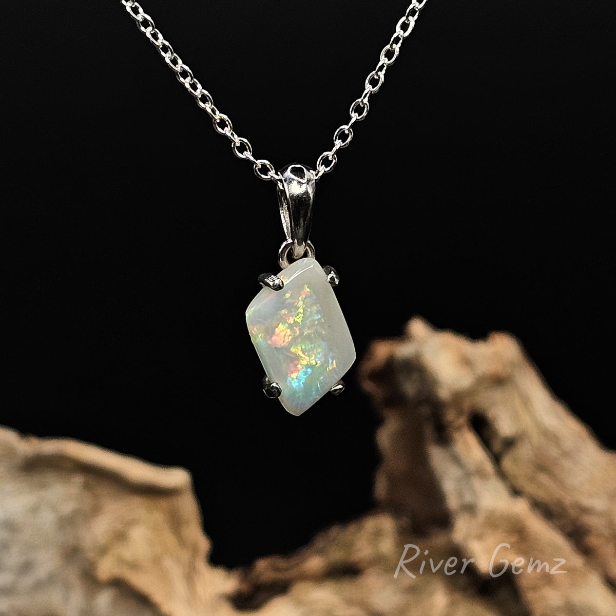 Fire like colours in light opal claw set in sterling silver pendant against a black background and light brown drift wood at the base with 'River Gemz' water mark in the bottom right hand corner.