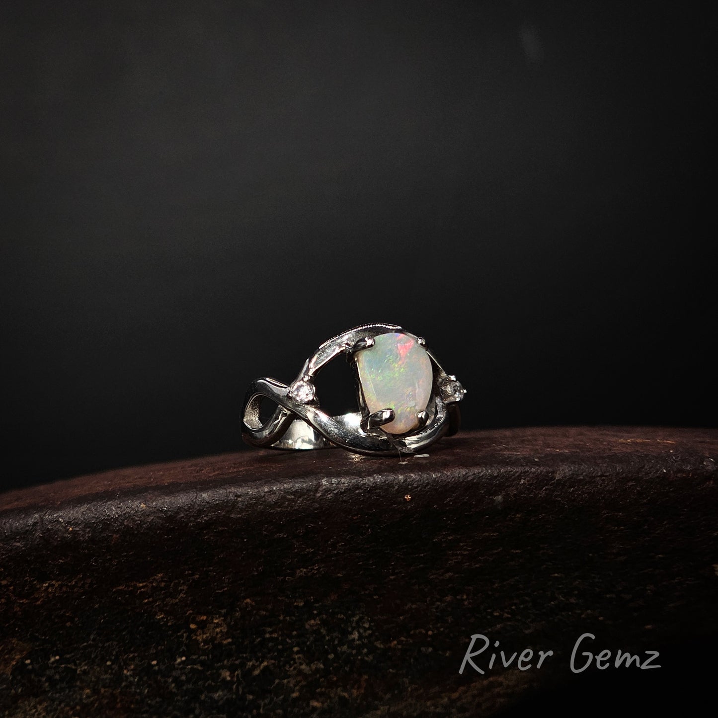 A silver ring featuring an oval iridescent white opal gemstone, displayed on a dark wooden surface against a dark background. The brand name 'River Gemz' is visible in the bottom right corner.