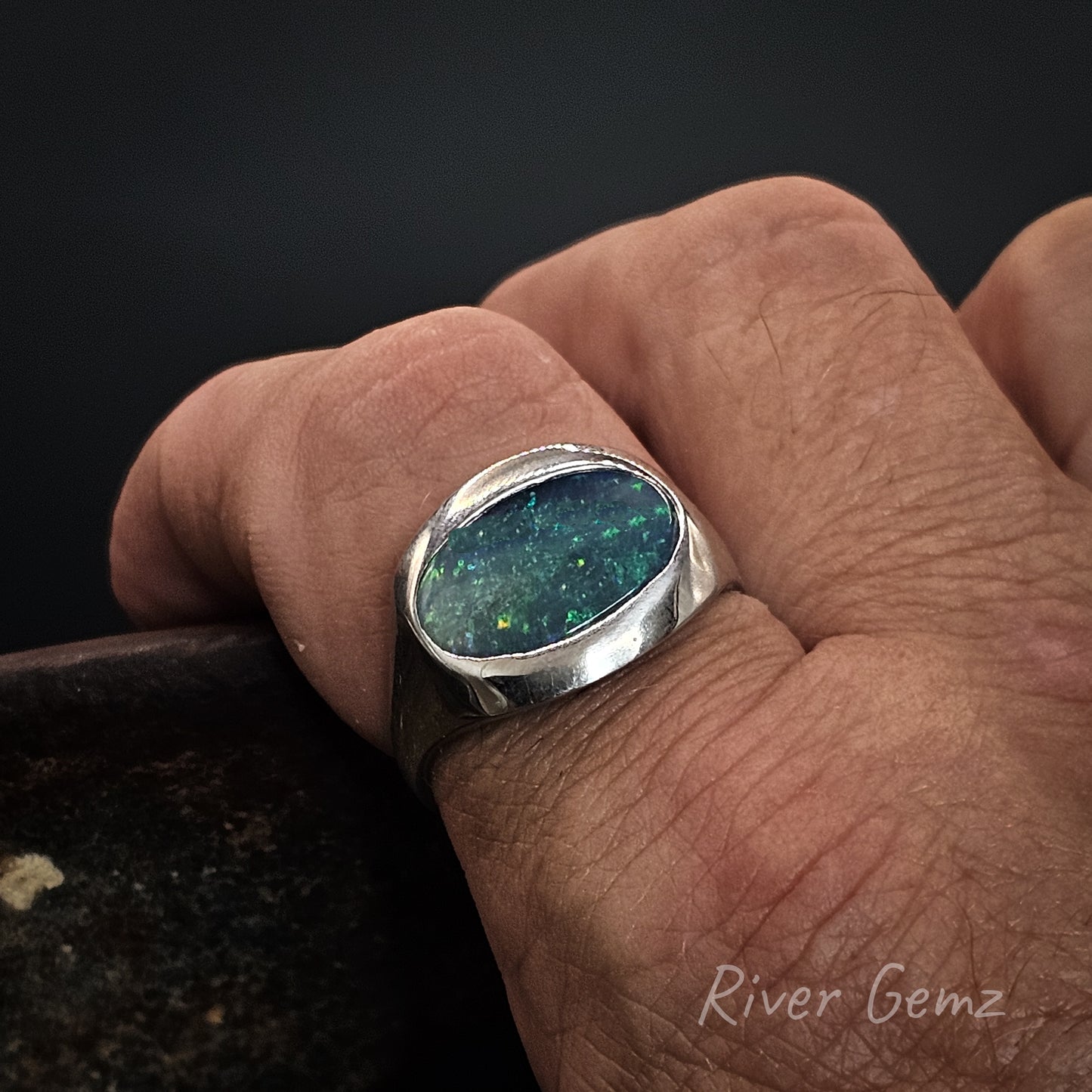 Hand wearing a silver ring with a blue-green stone, brand 'River Gemz' visible
