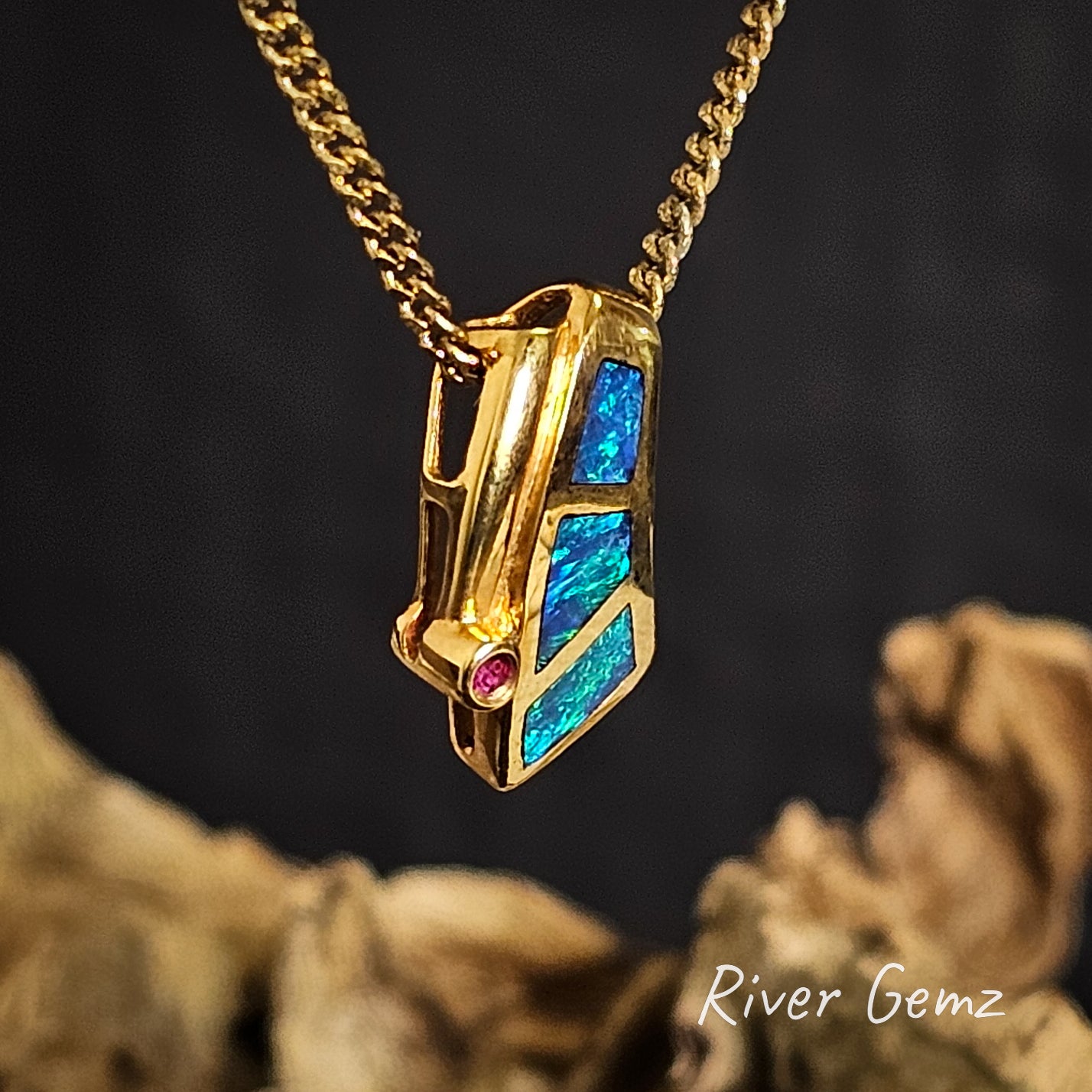 Gold necklace with a colorful pendant on a dark background branded 'River Gemz'.