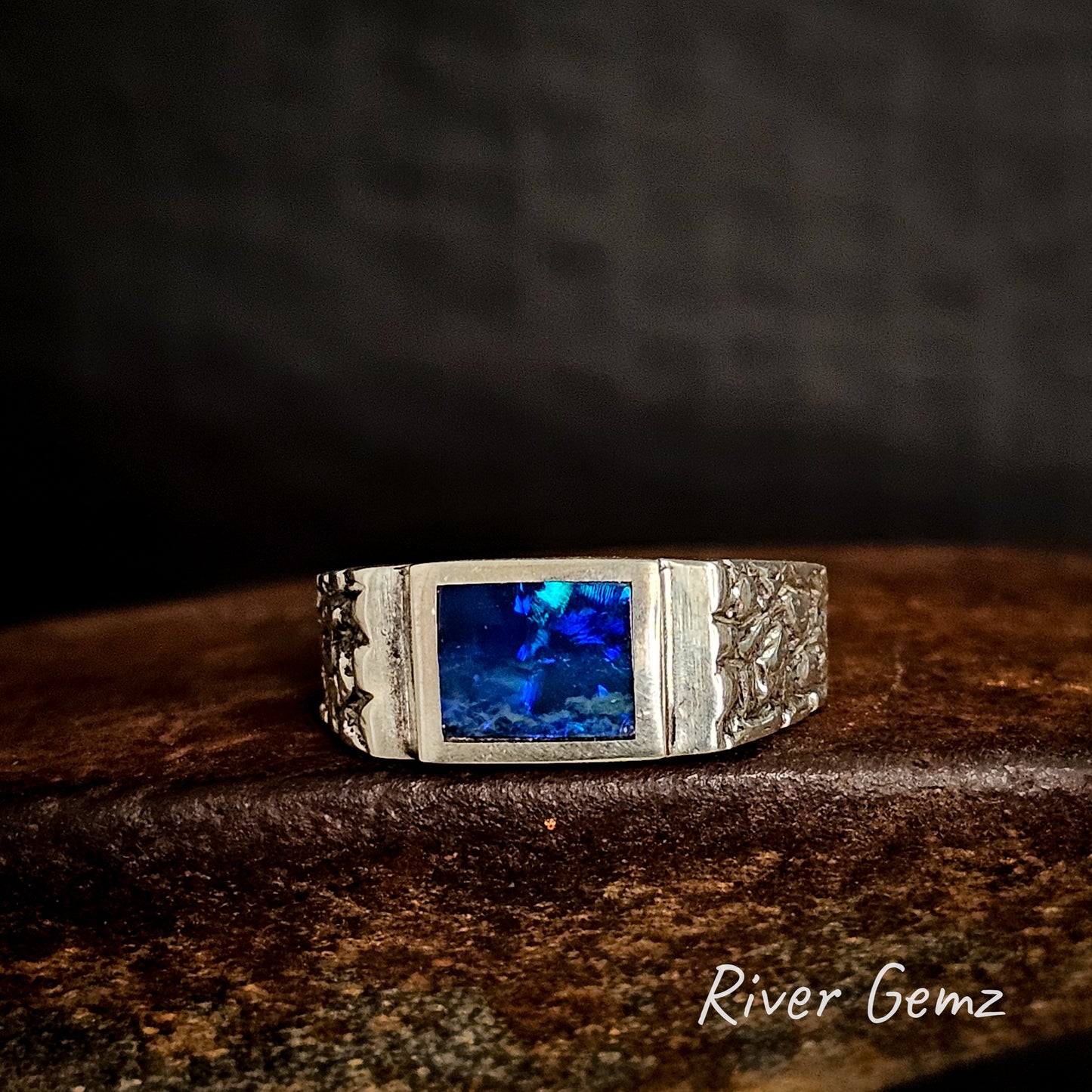 Silver ring with a blue gemstone on a dark background, featuring 'River Gemz' branding.