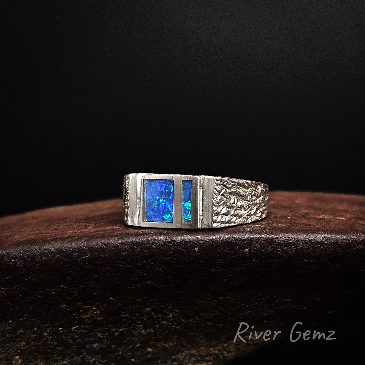 A silver ring featuring two rectangular blue opal gemstones, displayed on a dark wooden surface against a black background. The brand name 'River Gemz' is visible in the bottom right corner.