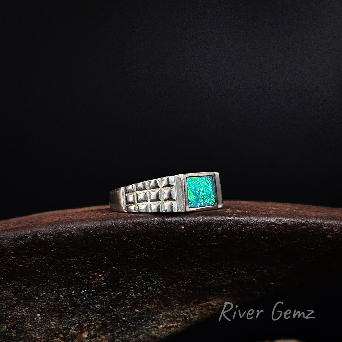 Vibrant green and blue rectangular opal inlaid in men's silver ring with checker plate tapered shoulders on a weathered shoe anvil and black background.