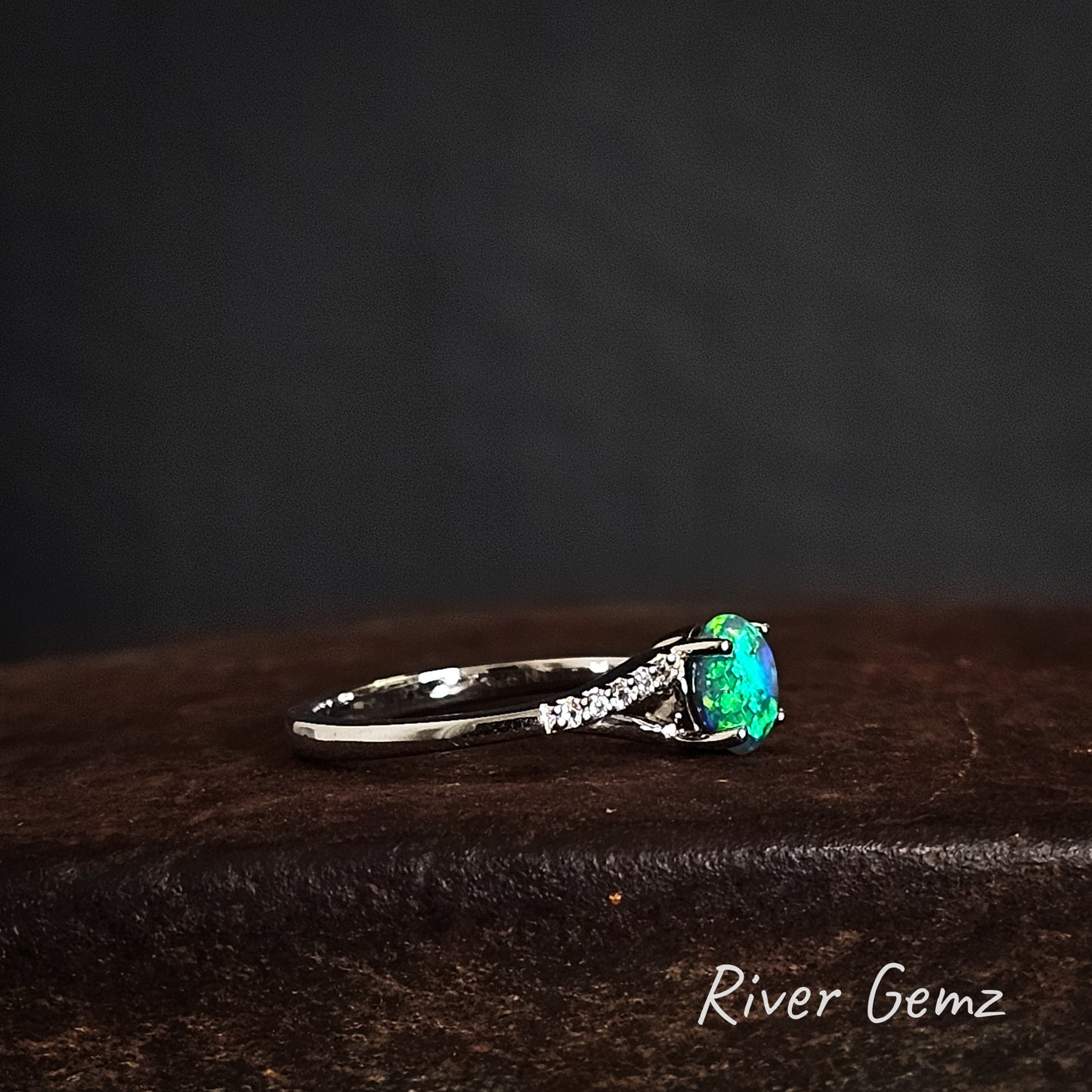 Side view of the silver ring with a bright green black opal and white topaz stones on the shoulder on a dark surface, branded 'River Gemz'.