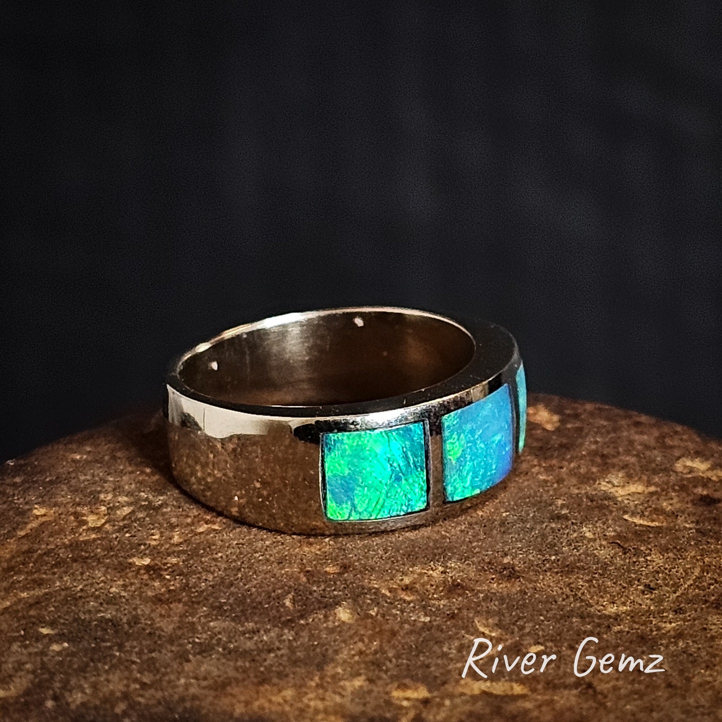 Side view of white gold ring with vivid green dark opals on a dark background, branded 'River Gemz'.