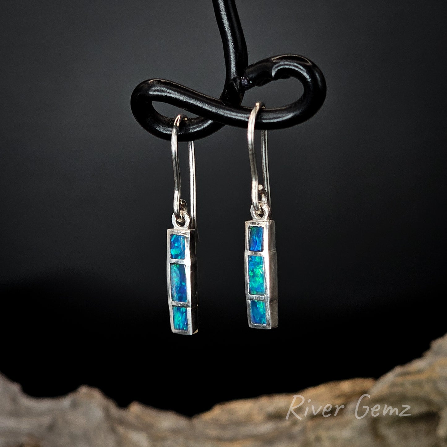 Pair of silver earrings with blue stones on a dark background, featuring 'River Gemz' branding.