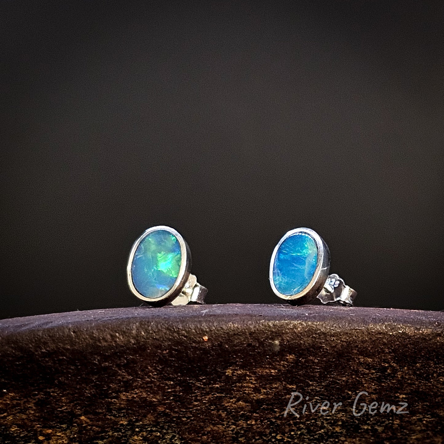 Pair of blue gemstone stud earrings on a dark background with 'River Gemz' branding.