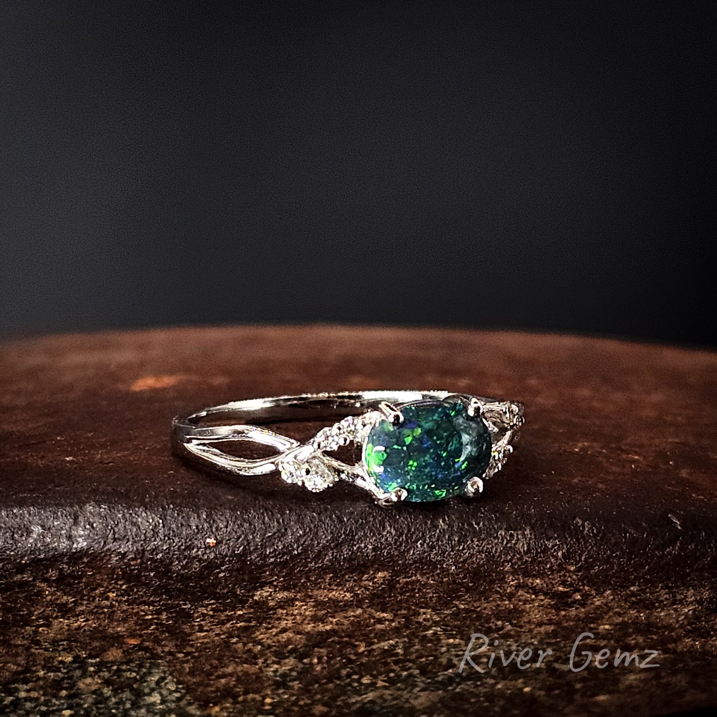 White gold ring with a green gemstone on a dark wooden surface, branded 'River Gemz'.
