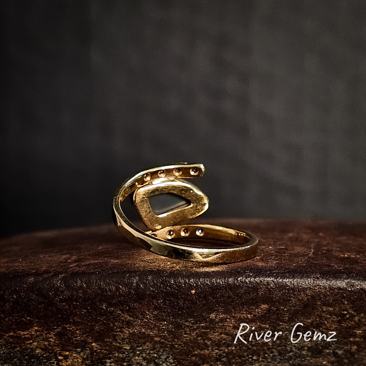 Rear view of the gold ring showing the dark tone of the black opal set in it on a dark surface with 'River Gemz' branding.