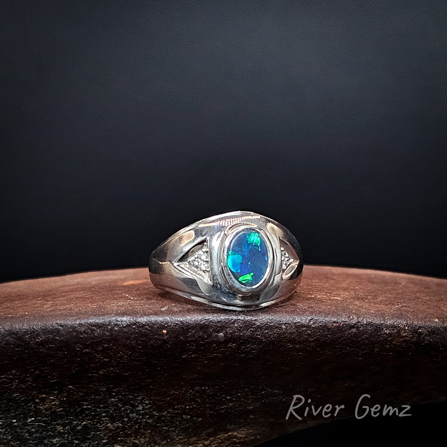 A sterling silver ring featuring a central oval blue opal gemstone, displayed on a dark wooden surface against a dark background. The brand name 'River Gemz' is visible as a watermark in the bottom right corner.