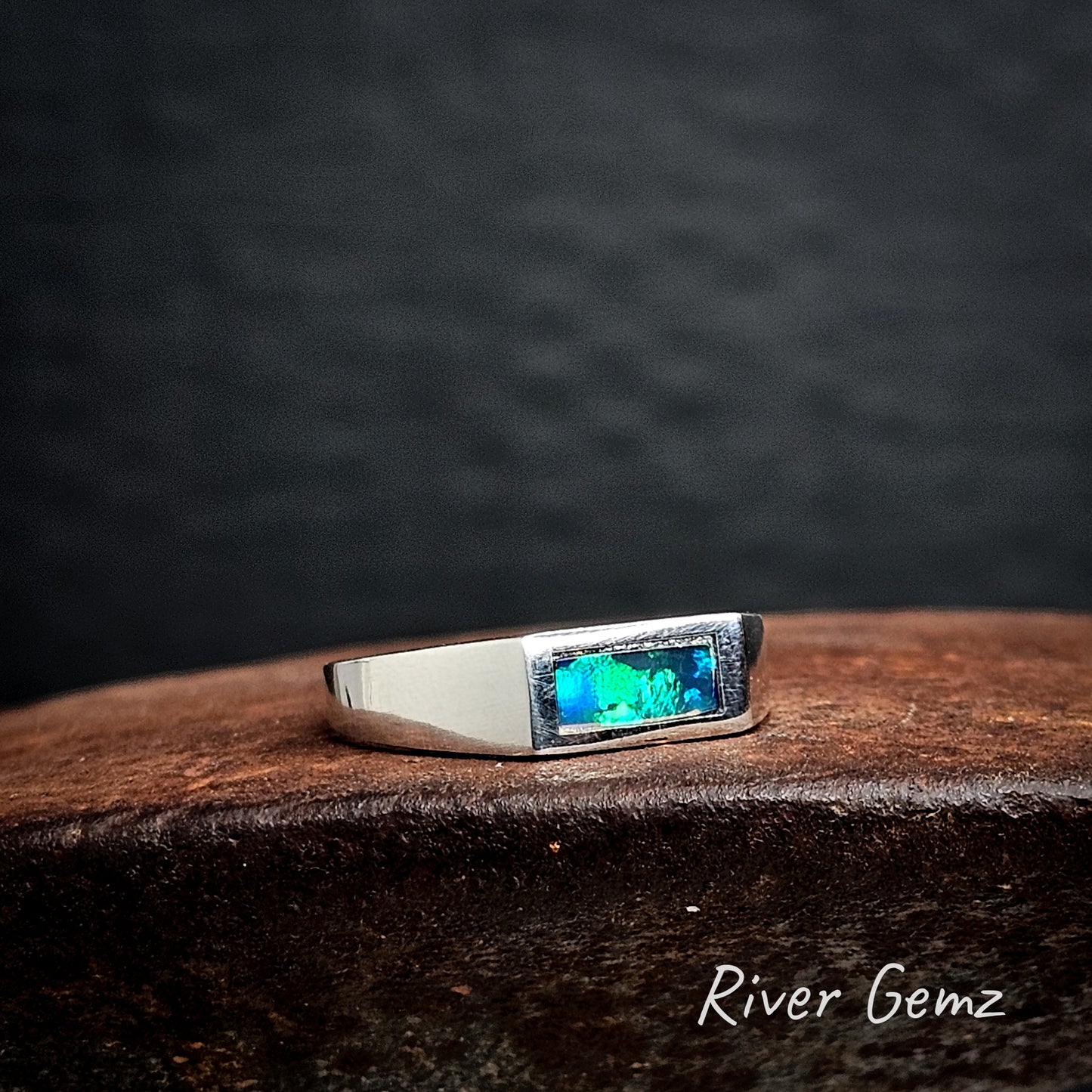 Silver ring with a blue and green gemstone on a cast iron surface, branded 'River Gemz'.