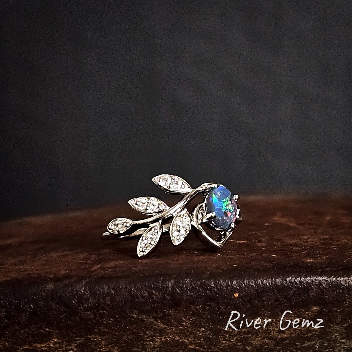 Silver leaf-shaped ring with a colourful black opal on a dark surface, branded 'River Gemz'.