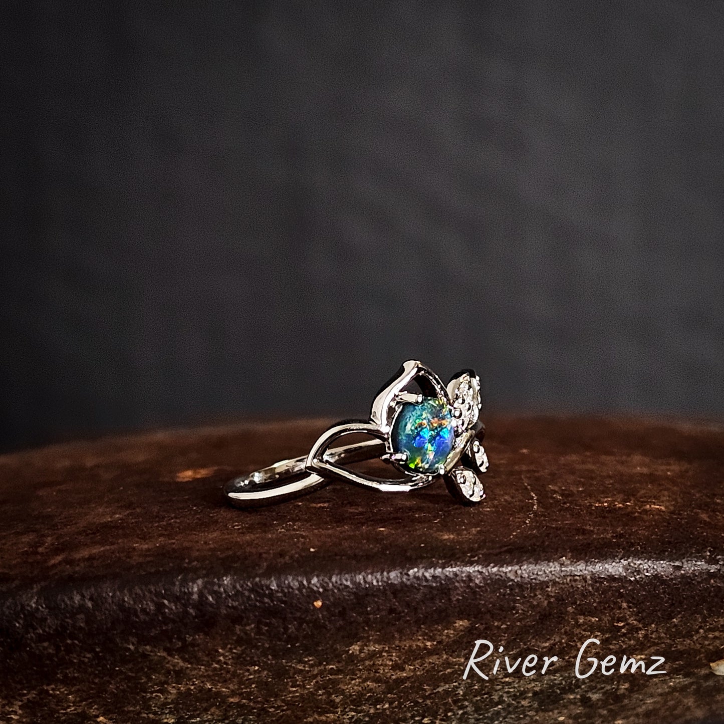 Silver ring with a colourful black opal on a dark brown surface, branded 'River Gemz'.