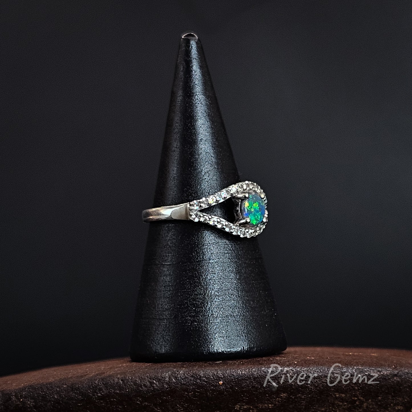 A silver ring with a vibrant green-blue opal center stone and a halo of small clear stones, displayed on a black cone stand against a dark background.
