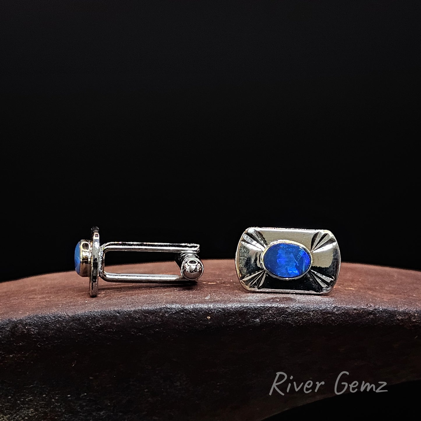 A pair of silver-toned cufflinks displayed on a dark brown wooden surface against a solid black background. One cufflink is shown from the side, revealing a blue oval gemstone, while the other is shown from the front with a textured silver bezel and the same blue gemstone.