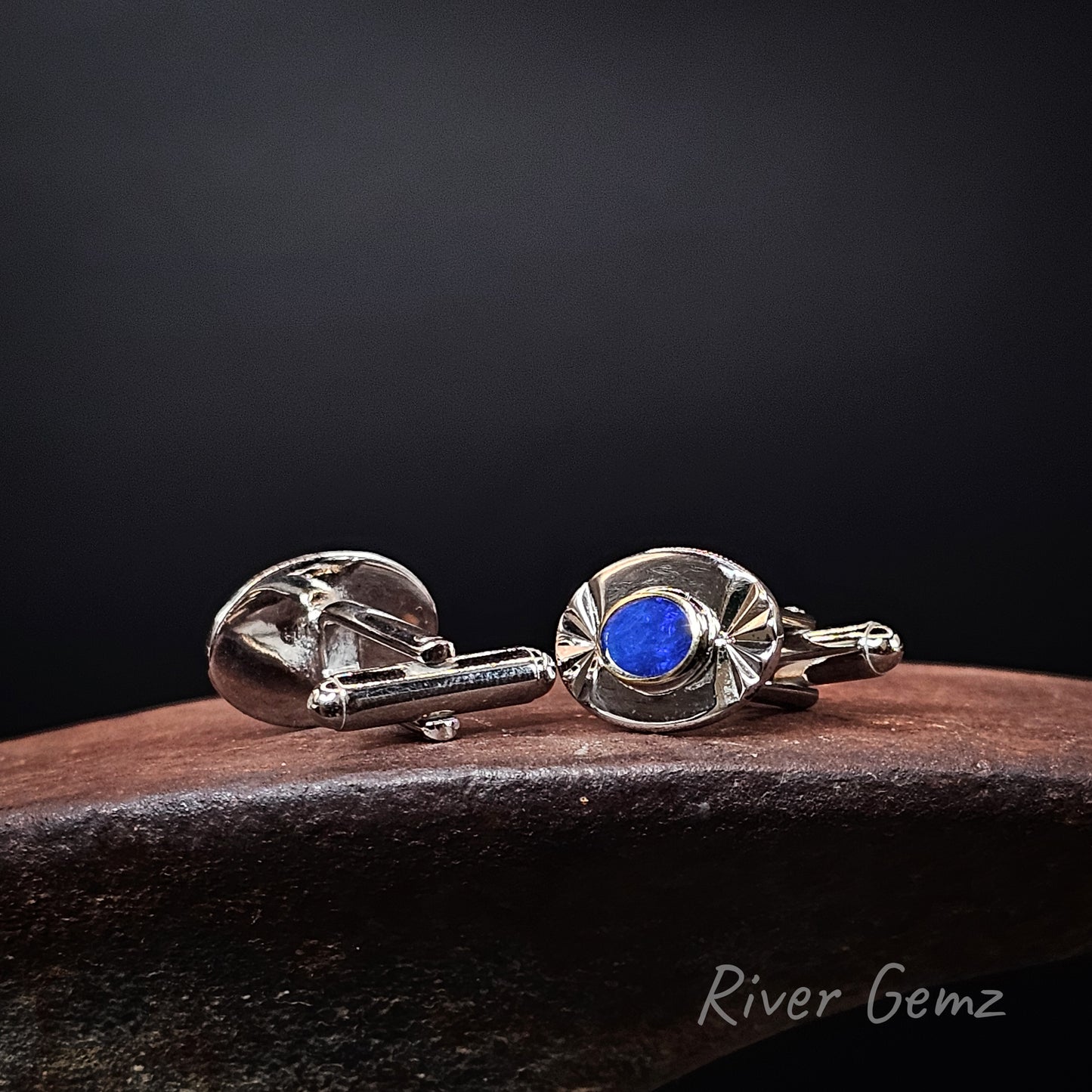 A pair of silver-toned cufflinks with oval blue gemstone accents, displayed on a dark wooden surface against a black background. The brand name 'River Gemz' is visible as a watermark in the bottom right corner.