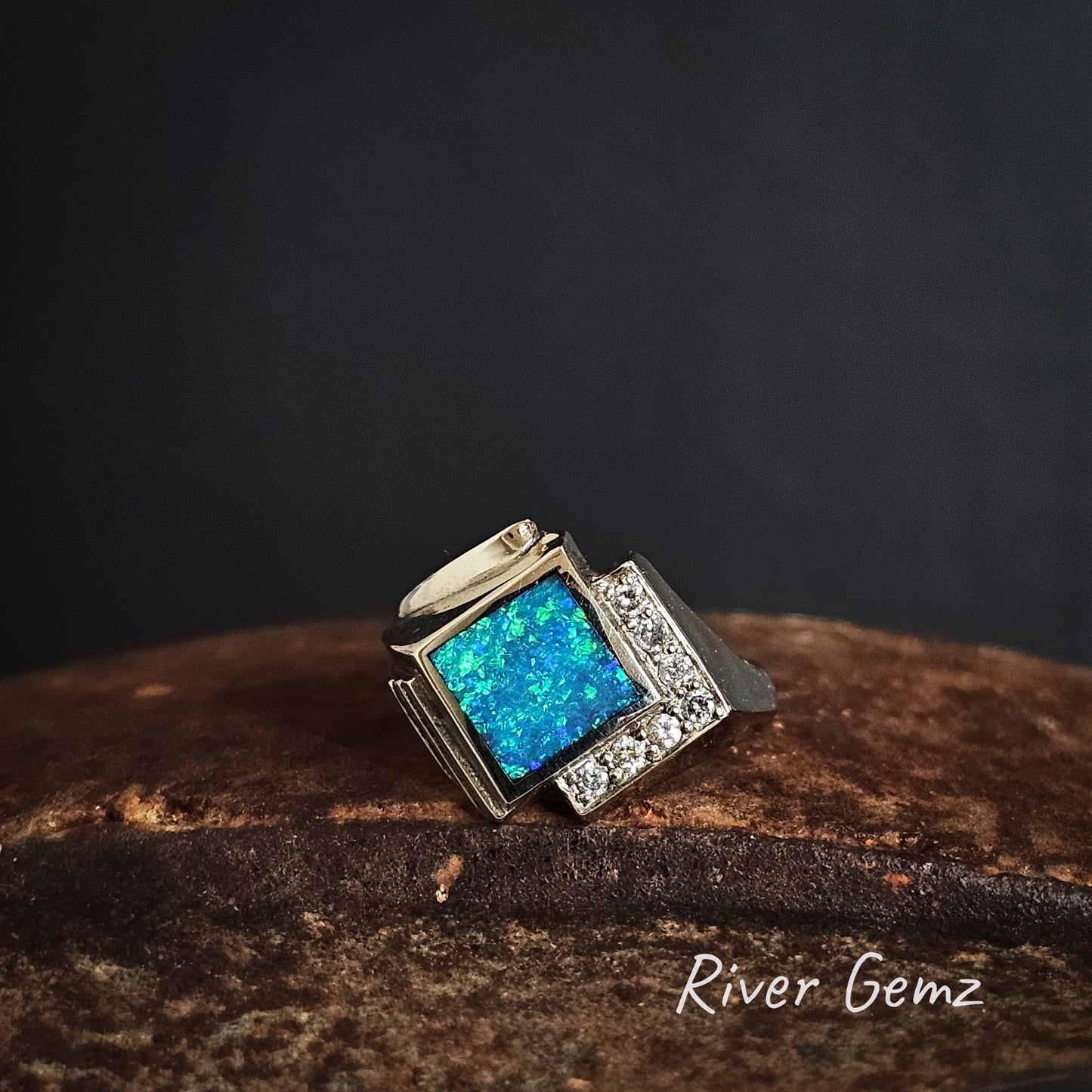 18K white gold ring with blue diamond shaped opal and diamonds on two sides on a dark background, featuring the brand 'River Gemz'.