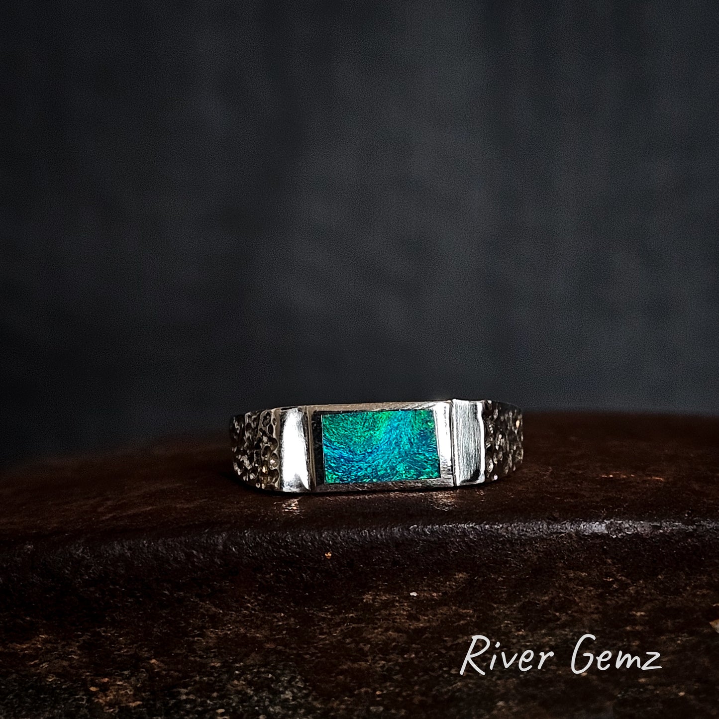 Silver ring with a blue-green gemstone on a dark background, branded 'River Gemz'.