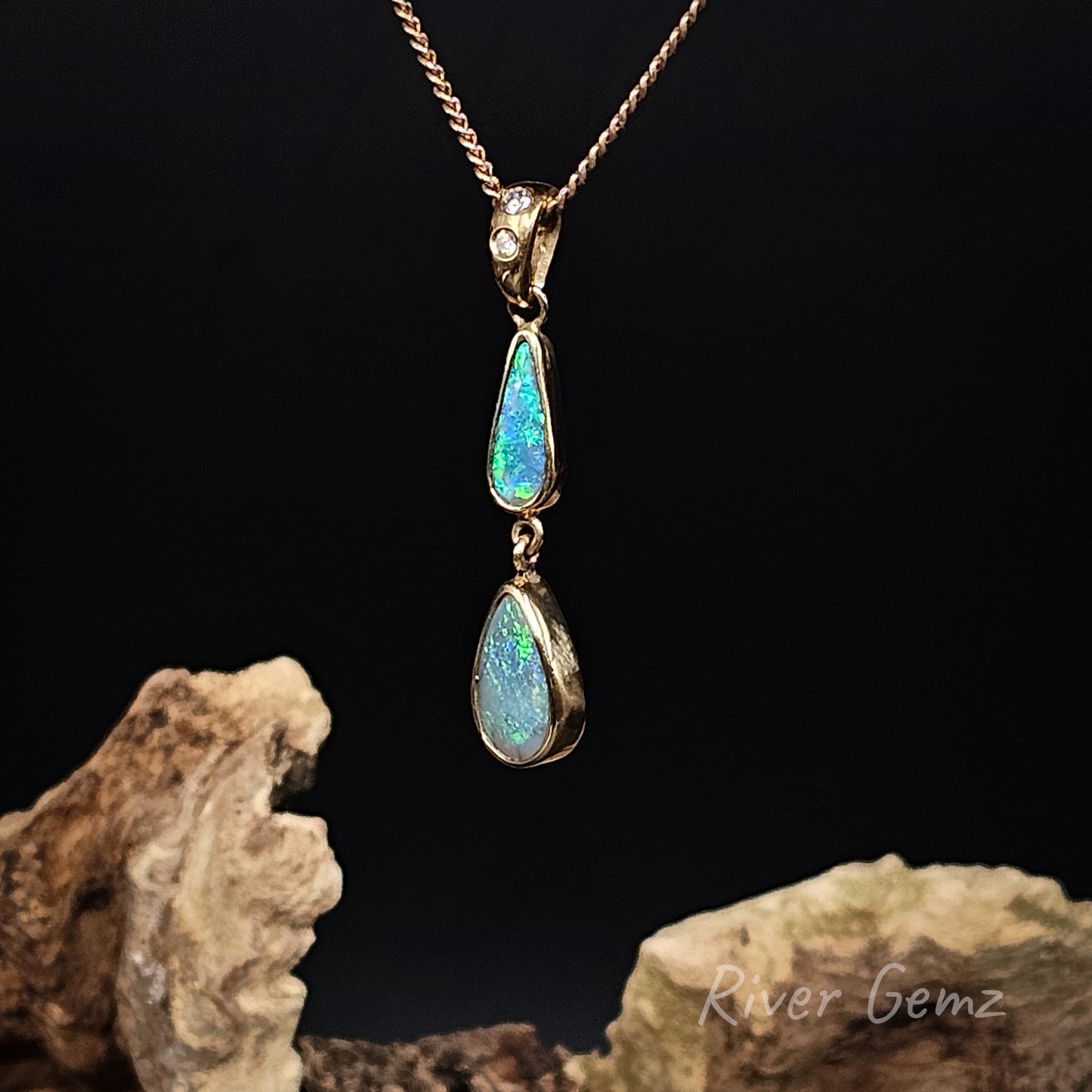A gold pendant necklace featuring two dangling teardrop-shaped iridescent opal gemstones, displayed against a black background with decorative driftwood props. The brand name 'River Gemz' is visible as a watermark in the bottom right corner.