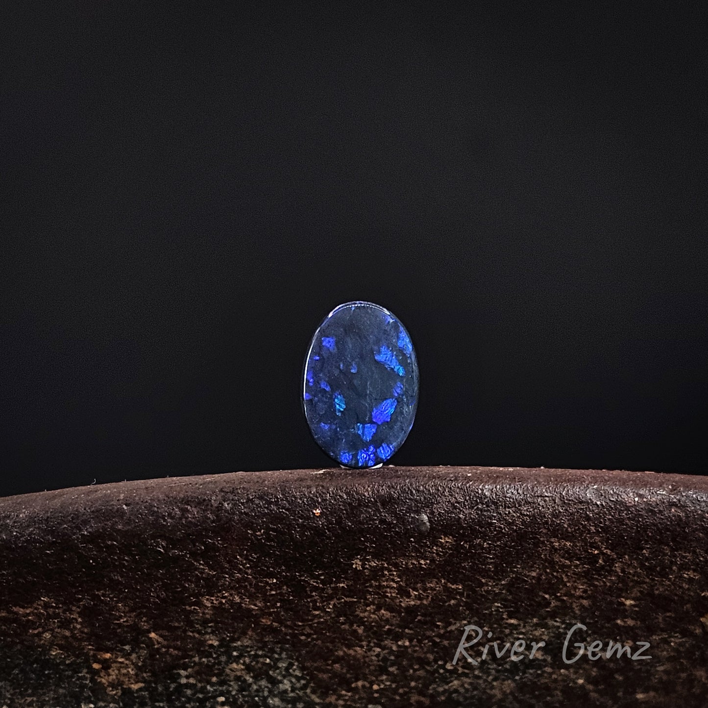 Unset dark opal with large spheres of dark blue, green and purple colours. The stone is standing upright on a shoe anvil with 'River Gemz' branding in the bottom right hand corner.