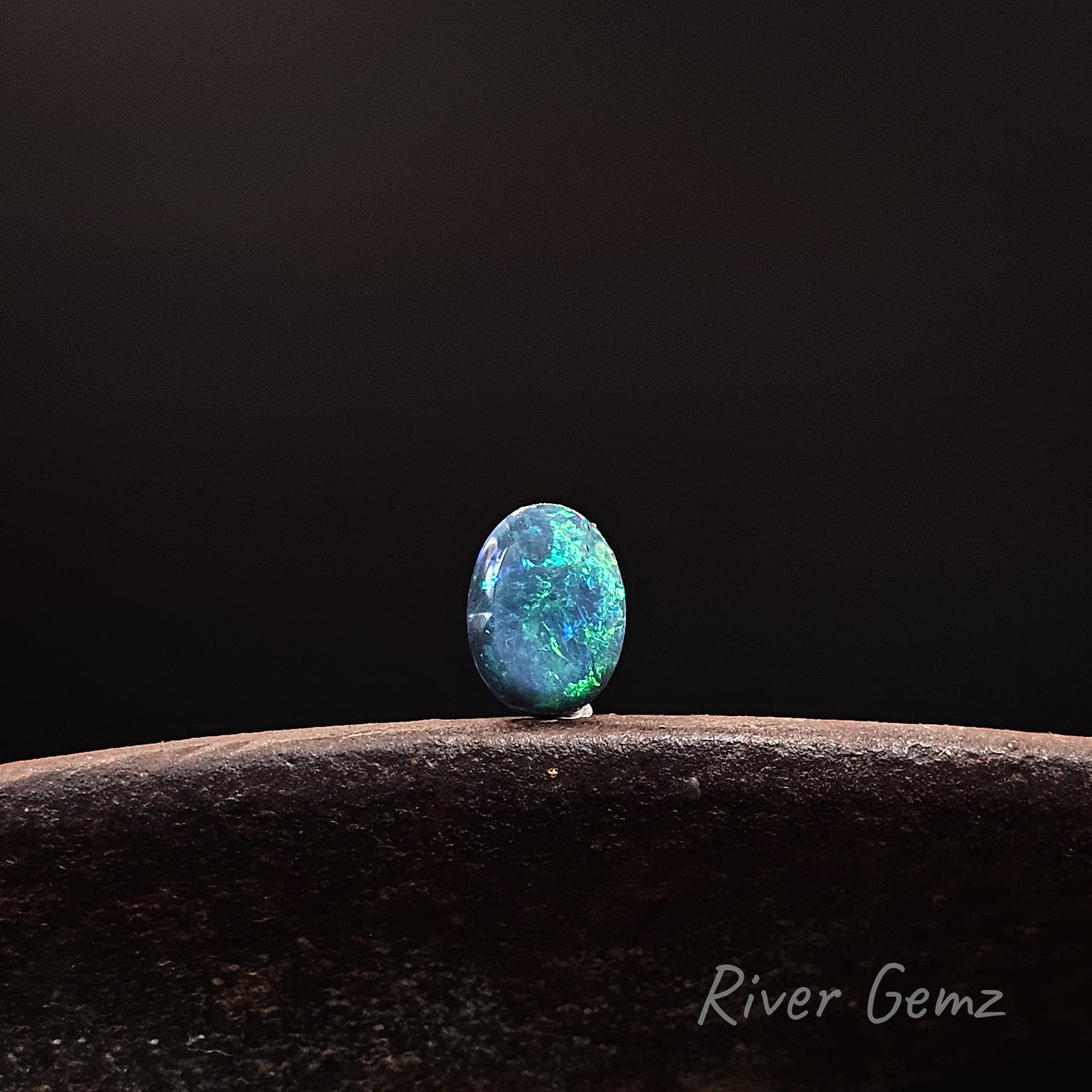 A single oval-cut blue opal gemstone with vibrant green and orange flashes, displayed on a dark textured stone surface against a solid black background. The watermark 'River Gemz' is visible in the bottom right corner.