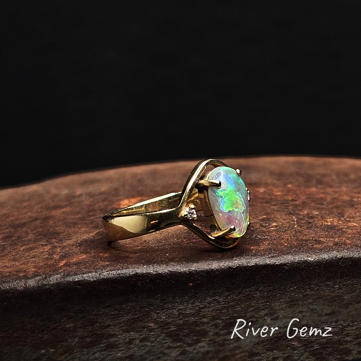 Gold ring with a colorful gemstone on a dark surface, branded 'River Gemz'.