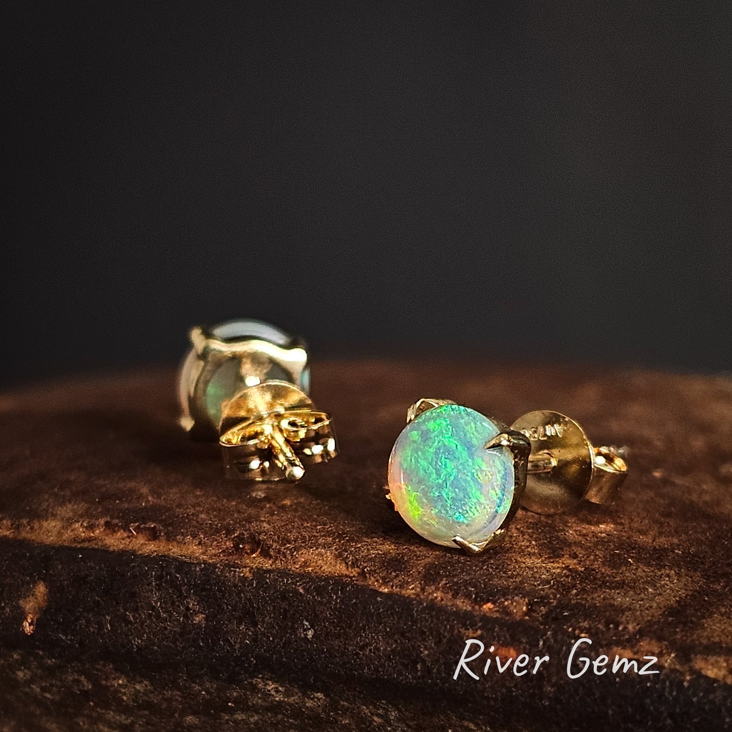 Opal stud earrings with yellow gold posts on a dark brown surface, branded 'River Gemz'.