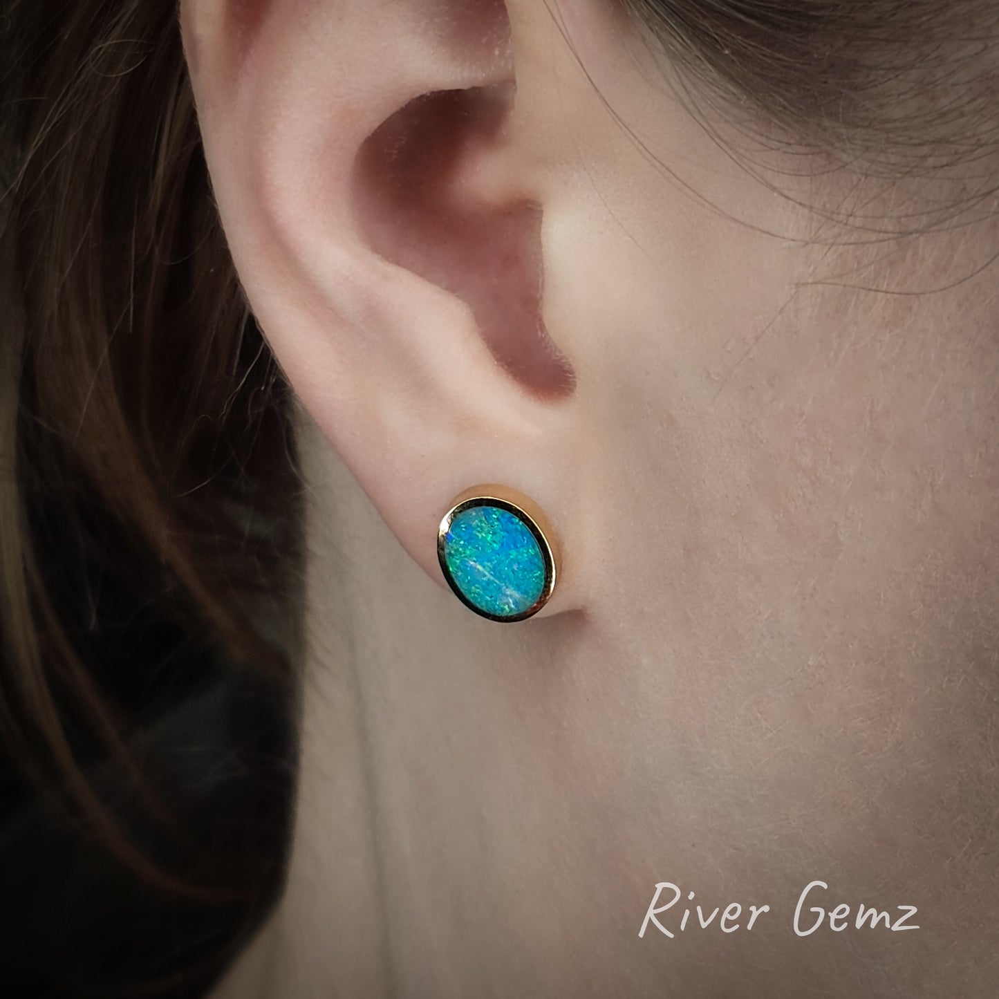 Oval shaped blue and green opal besel set in yellow gold stud in person's ear.