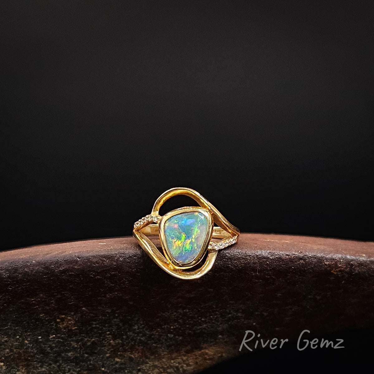 Yellow gold ring with central opal and white diamonds on the shoulders on a weathered shoe anvil, black background and 'River Gemz' brand in the bottom corner.