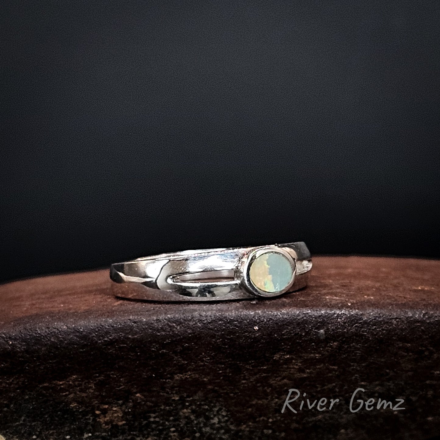 Silver ring with an oval shaped dark opal on a brown coloured base with 'River Gemz' branding.