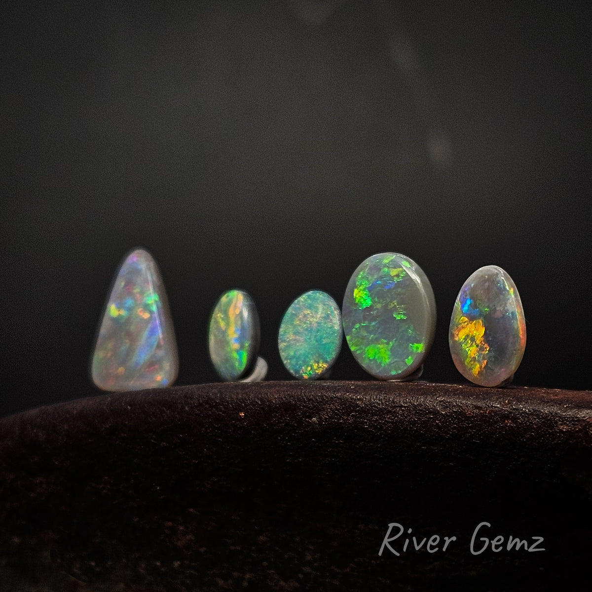 5 unset multi-coloured opals standing upright on an shoe anvil with 'River Gemz' watermark in the bottom right hand corner.