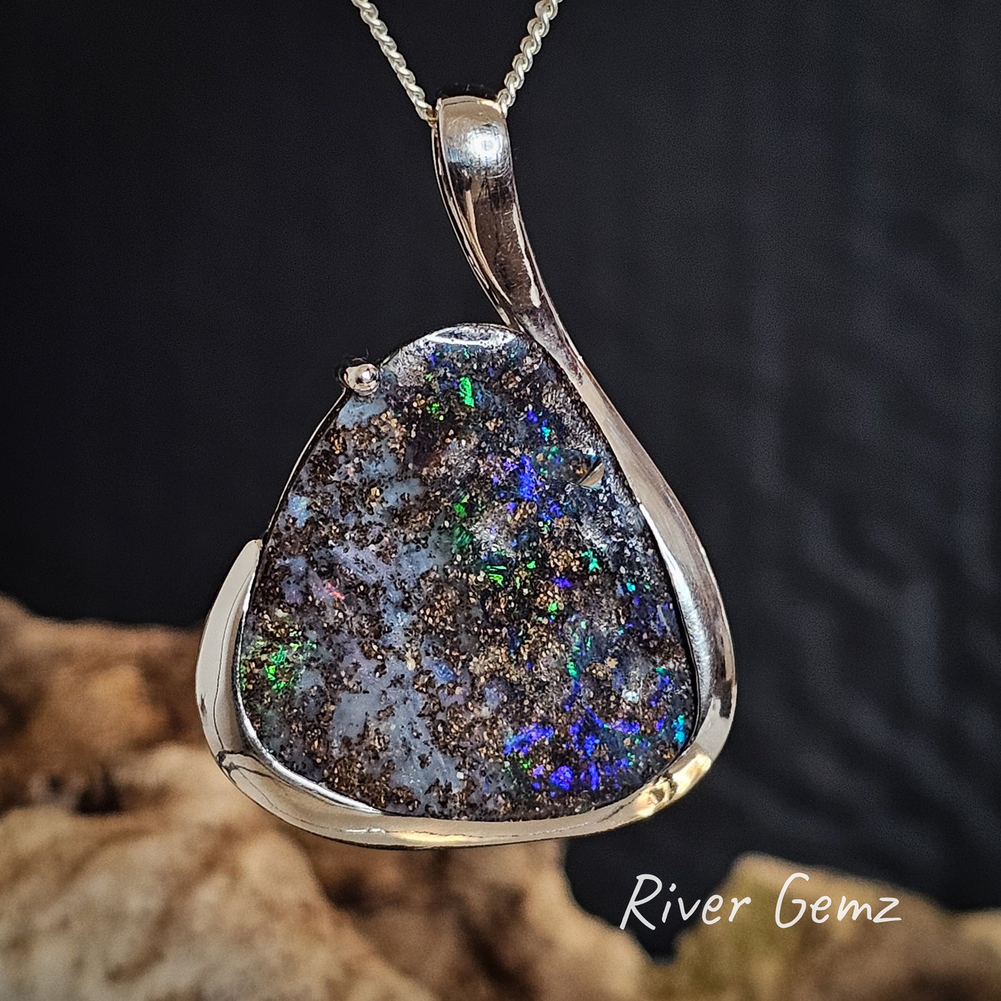 Teardrop-shaped pendant with multicolored gemstone on a dark background, branded 'River Gemz'.
