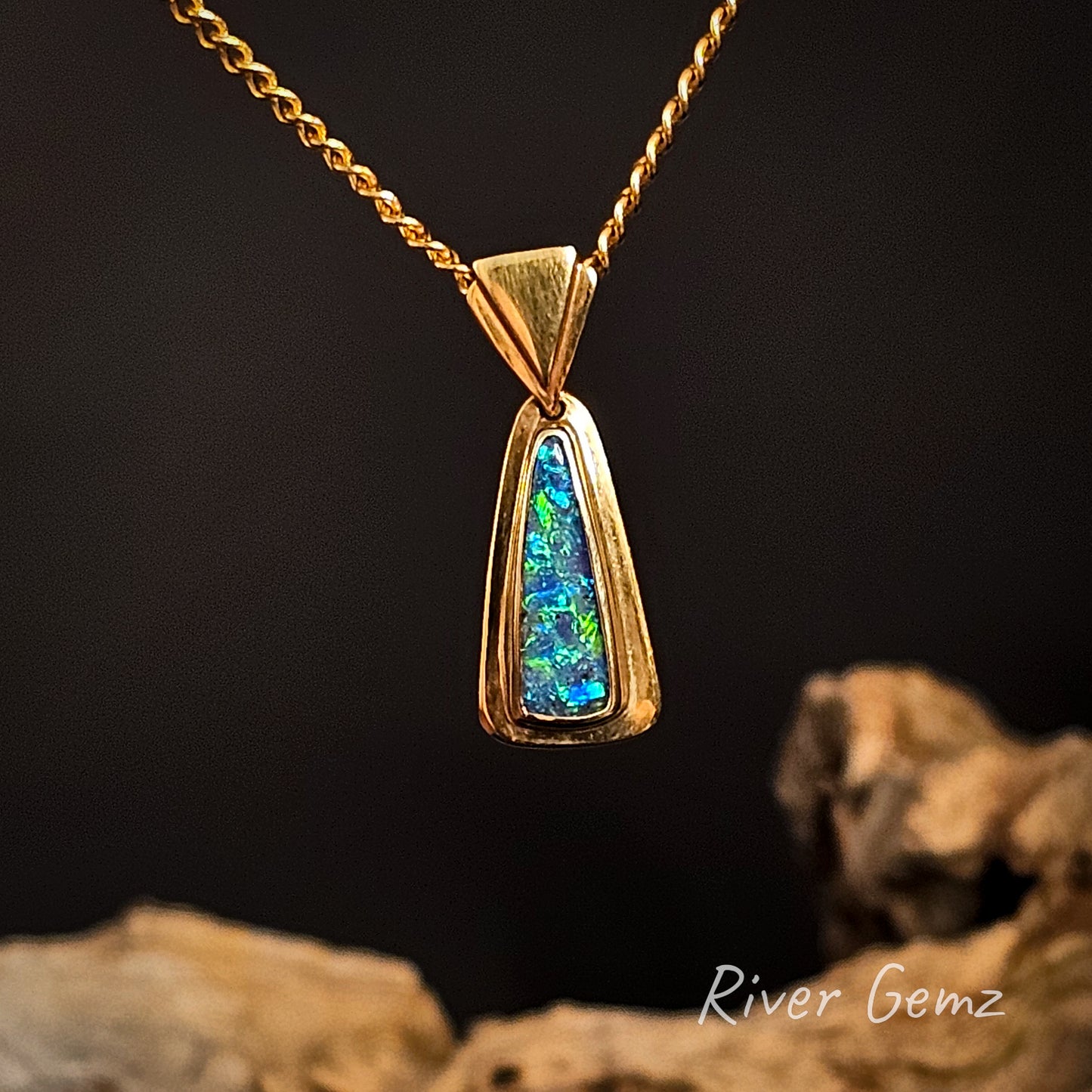 Gold pendant with a colorful gemstone on a dark background, featuring 'River Gemz' branding.