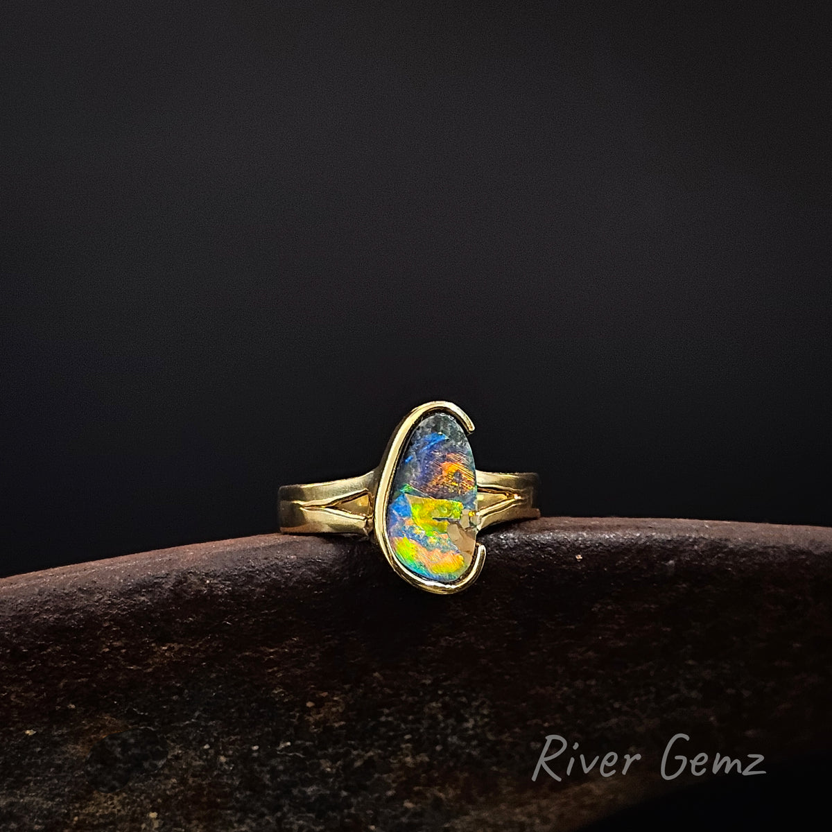 Gold ring with vibrantly coloured opal on a weathered shoe anvil against a dark background and 'River Gemz' watermark in the bottom right hand corner.
