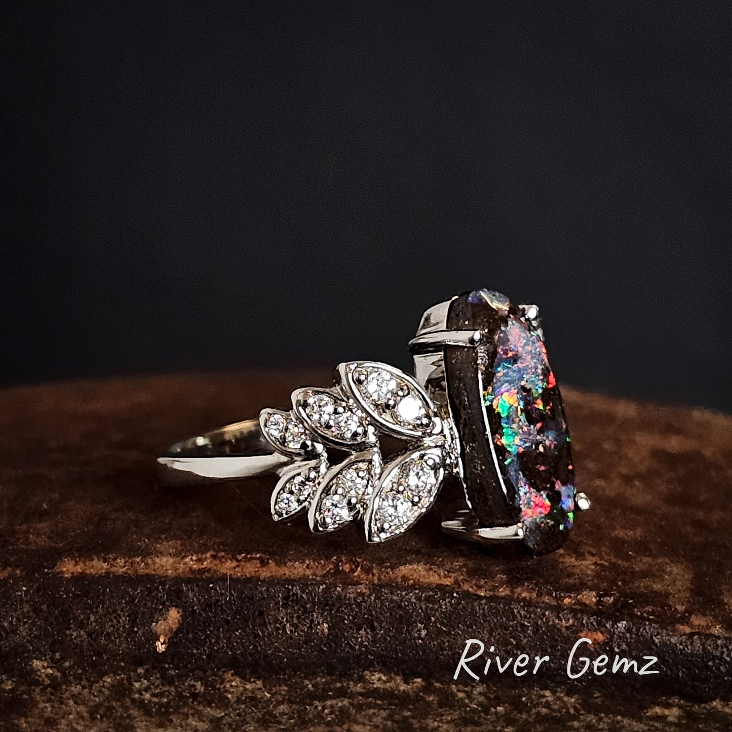 Side view of the ring showing the ironstone matrix of the boulder opal and the fern leaf with embedded diamonds on a dark background with 'River Gemz' branding.