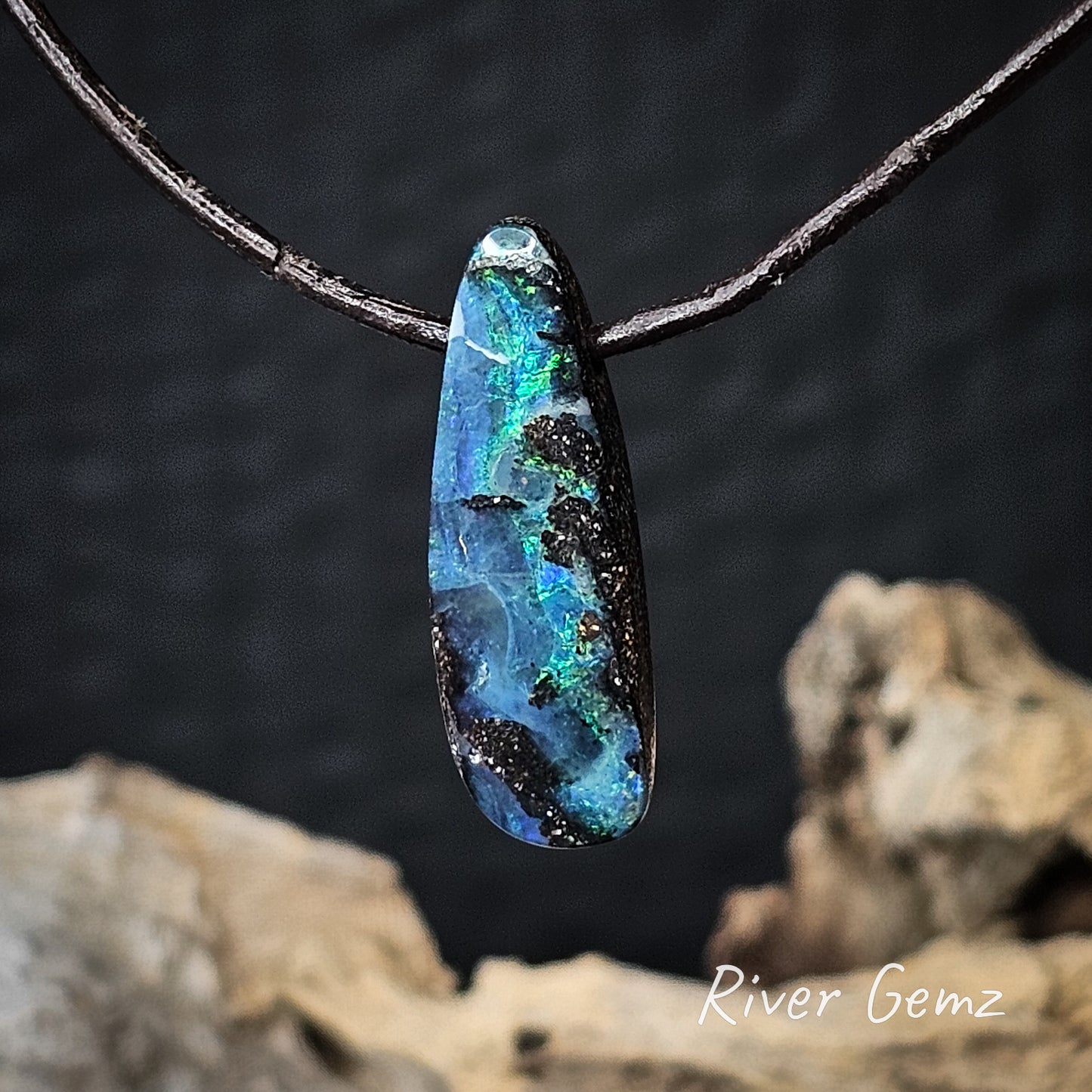 Opal pendant on a dark brown cord with a natural wood background, featuring the brand 'River Gemz'.