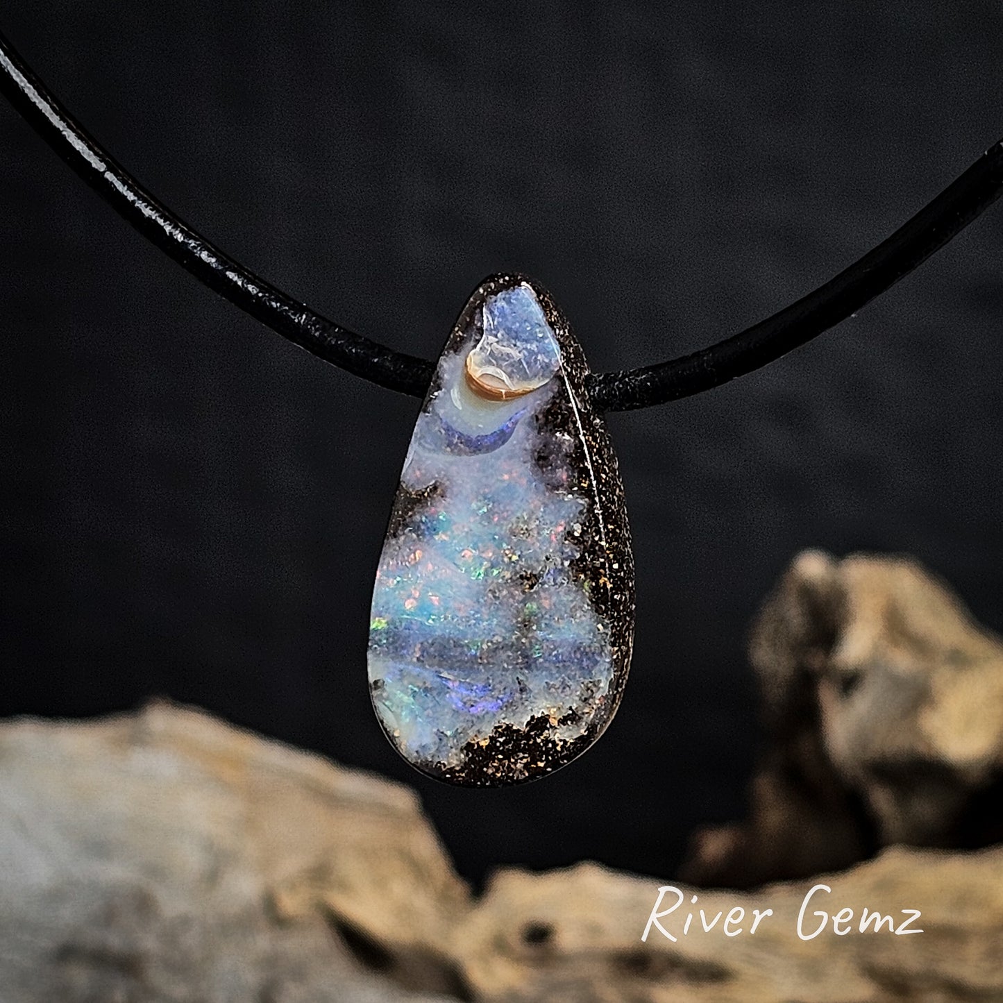 Multi-coloured teardrop-shaped opal pendant on a black cord with a blurred natural background.