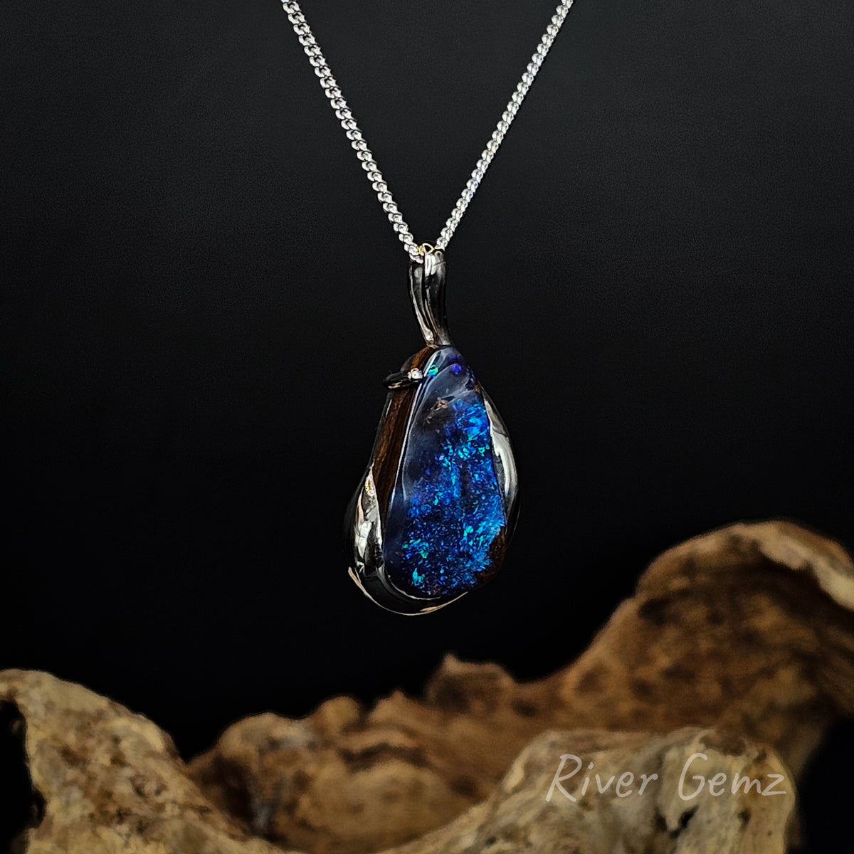 A teardrop-shaped pendant necklace with a vibrant blue boulder opal in a polished silver bezel setting, hanging from a silver chain against a dark background with a piece of driftwood at the bottom.