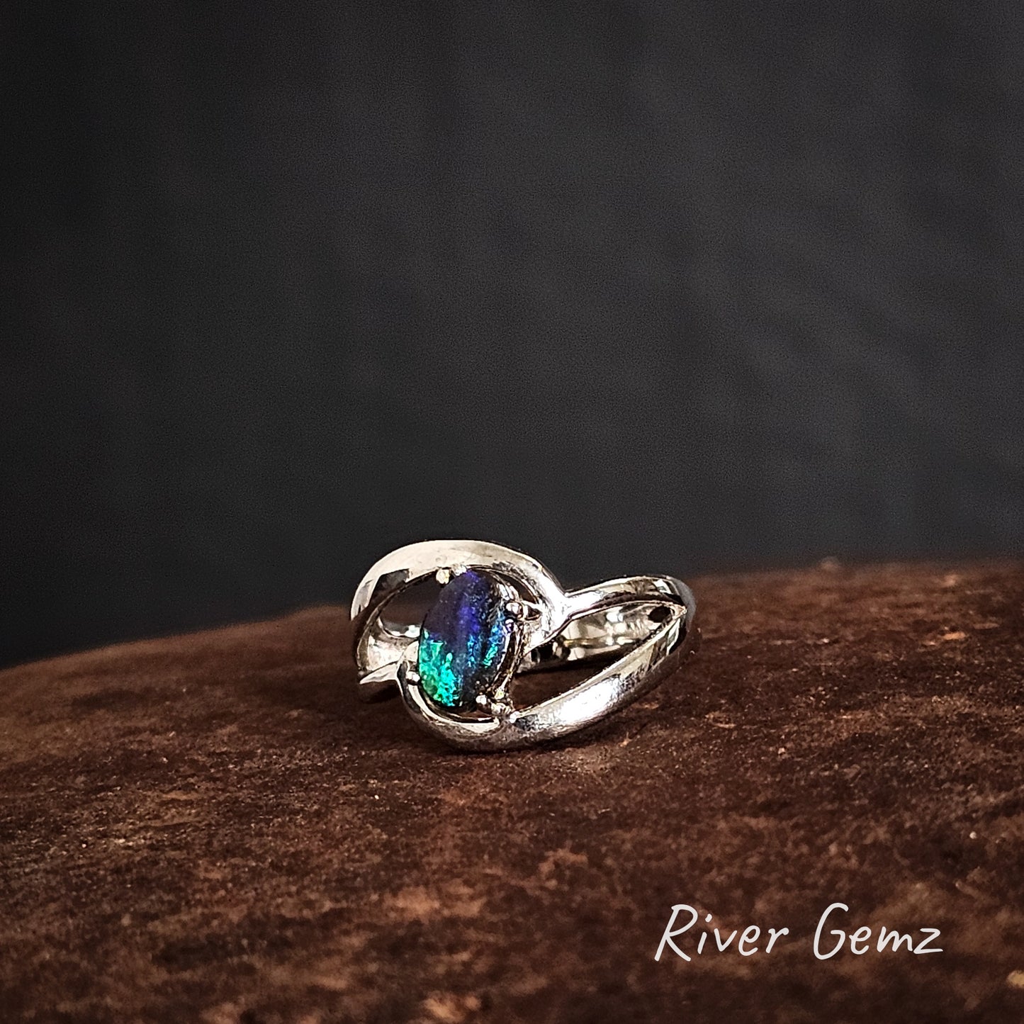 Silver ring with a colorful gemstone on a dark background, featuring the brand 'River Gemz'.