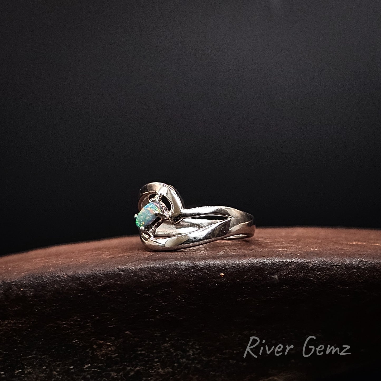 A silver ring with a curved, textured band and a small green opal gemstone, displayed on a dark brown wooden surface against a black background. The brand name 'River Gemz' is visible in the bottom right corner.