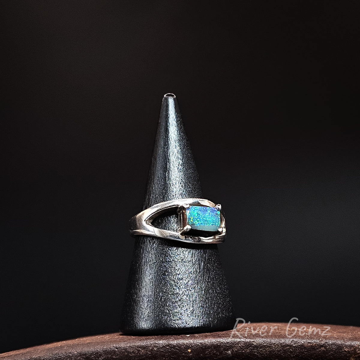 A silver ring with a rectangular blue-green opal gemstone, displayed on a black cone stand against a dark background.