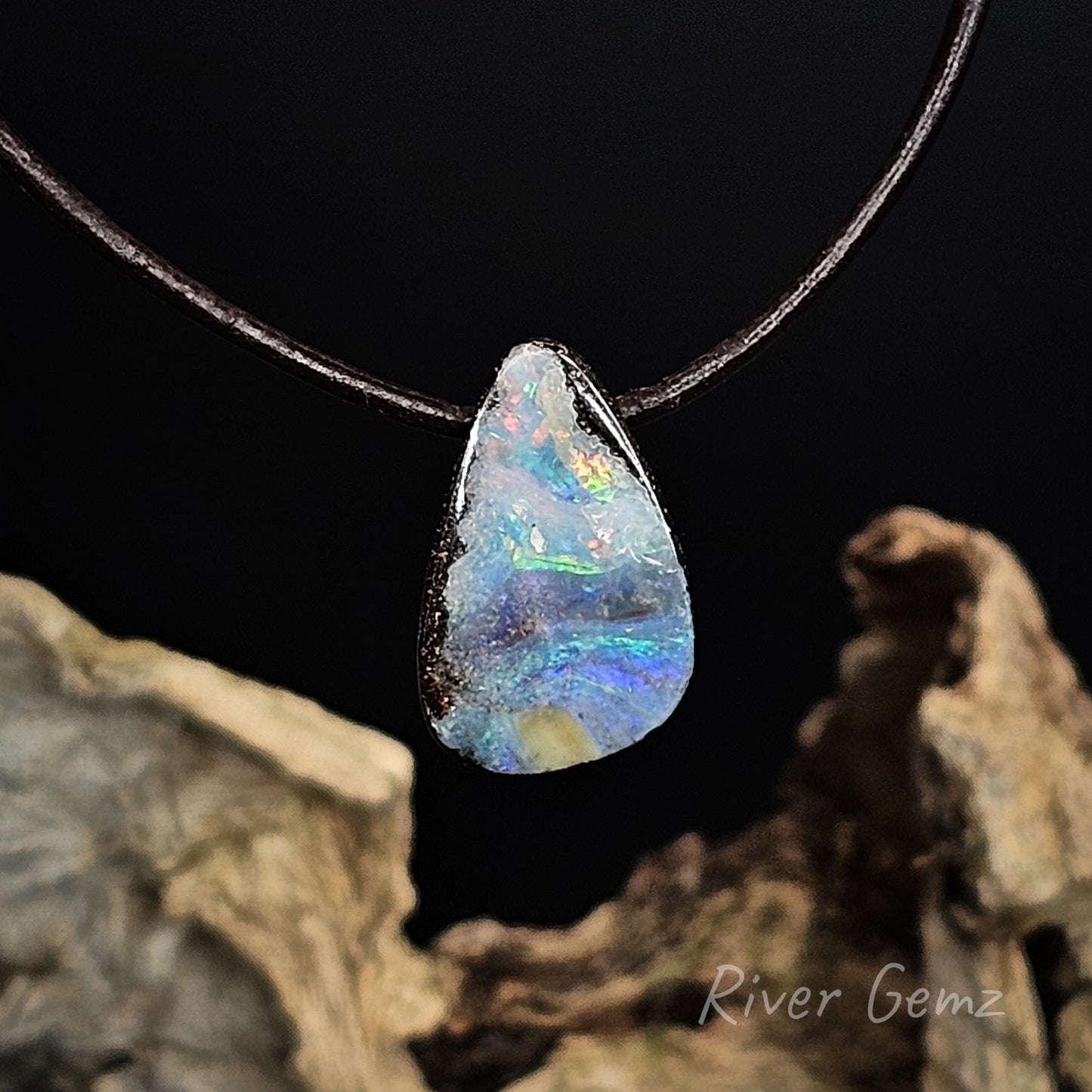 Opal pendant on a black cord with a natural rock background, featuring the brand 'River Gemz'.