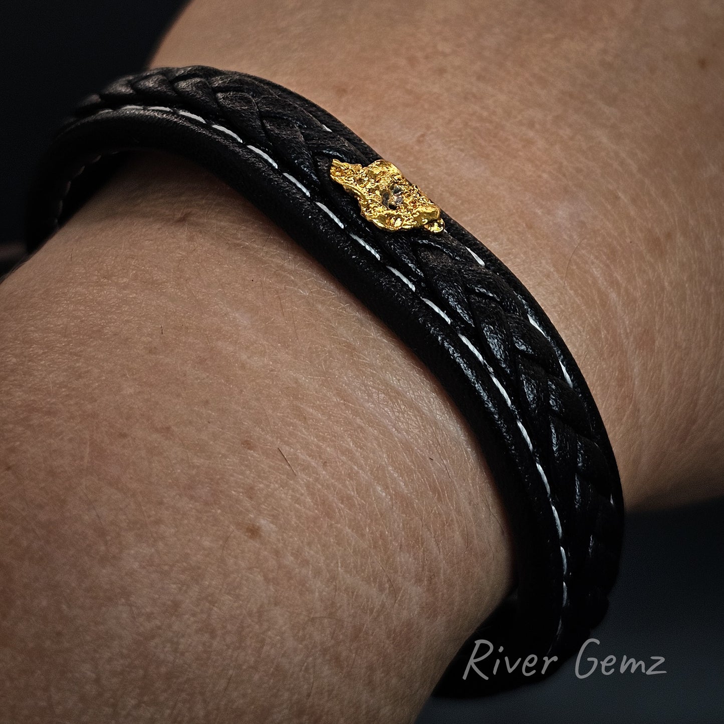 Black braided leather bracelet with a gold clasp on a wrist, branded 'River Gemz'.