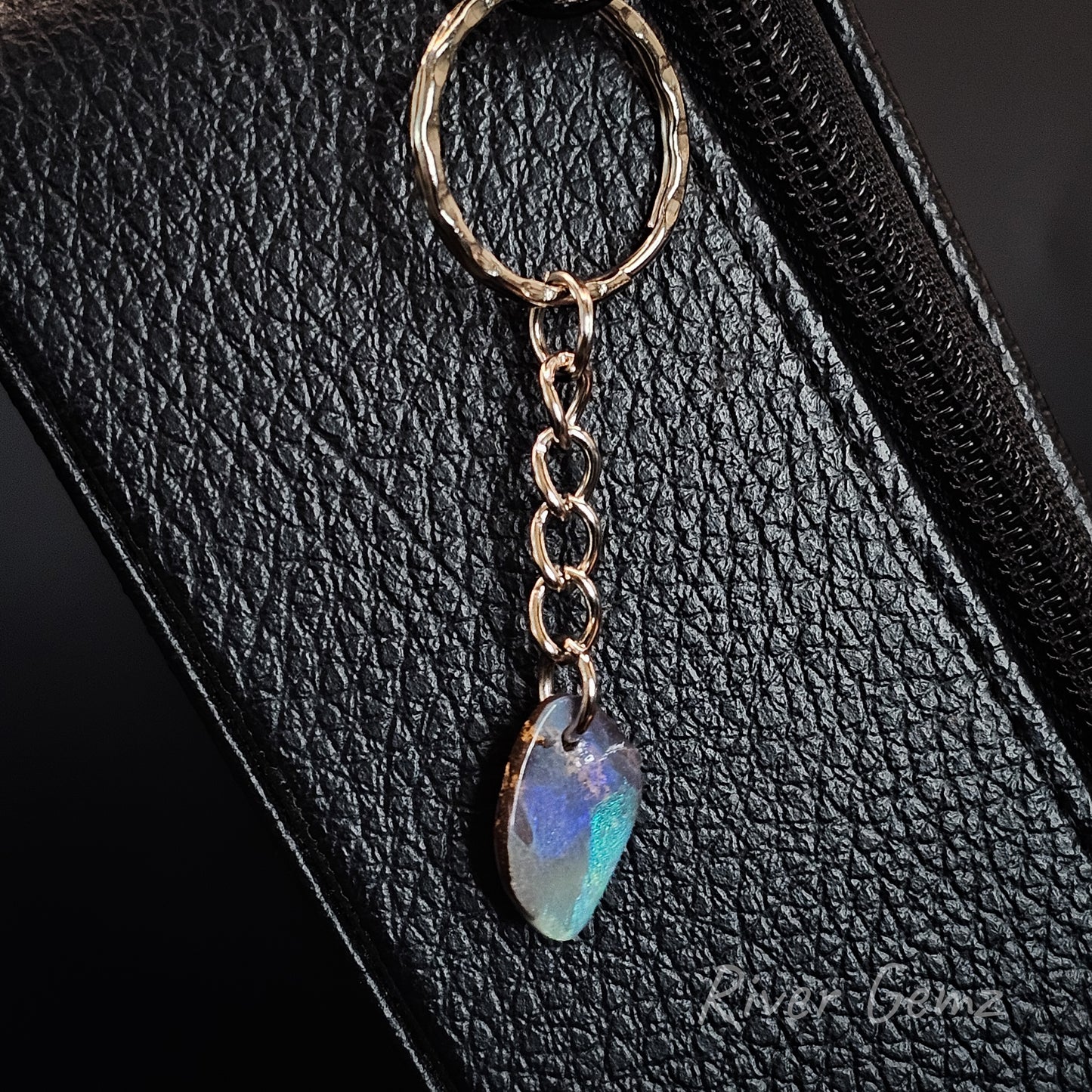 A close-up of a teardrop-shaped iridescent opal bag charm, resting on a black textured leather surface.