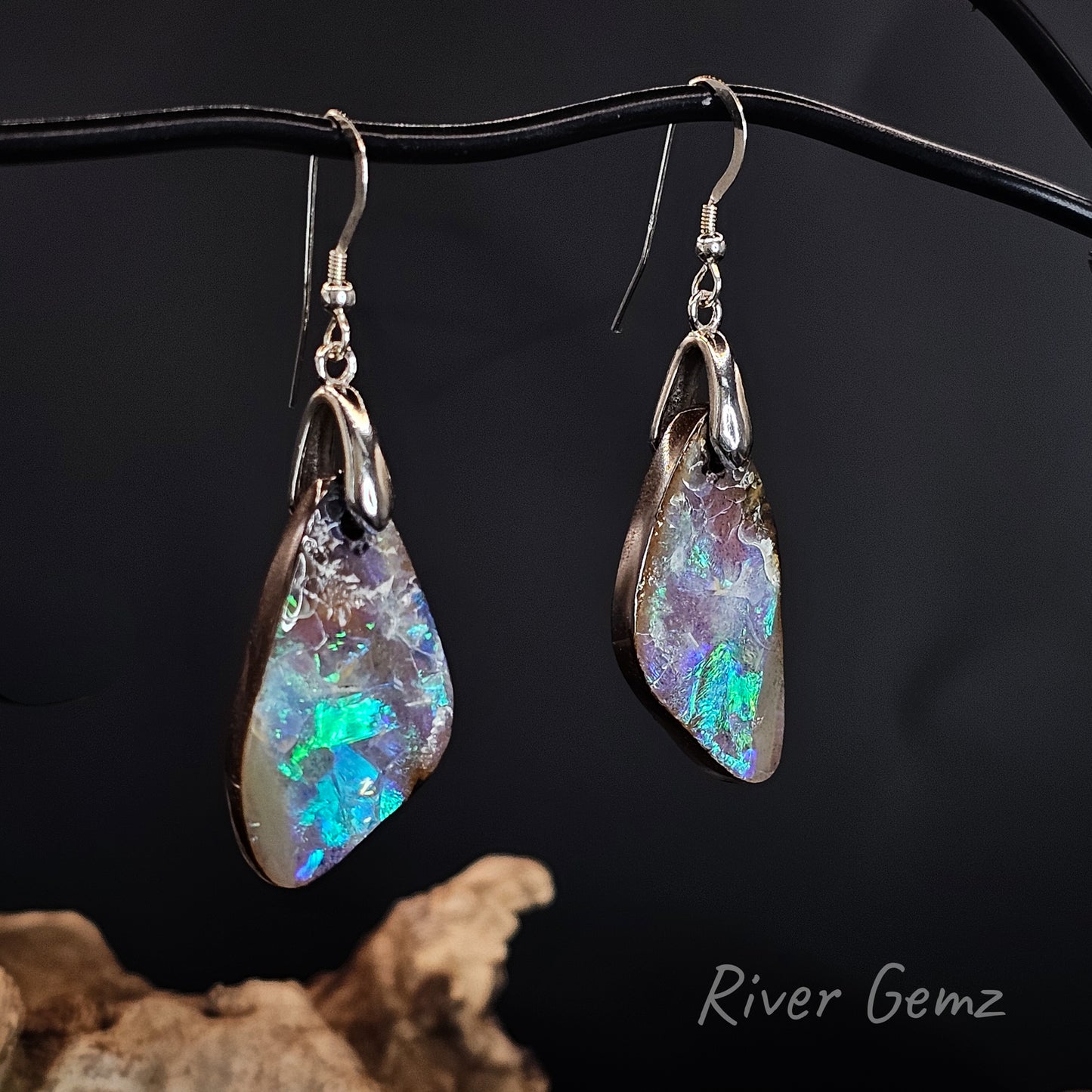 A pair of irregular teardrop-shaped opal dangle earrings with silver-toned metal settings and hooks, hanging against a black background with a piece of driftwood at the bottom. The opal stones show vibrant blue, green, and purple iridescence.