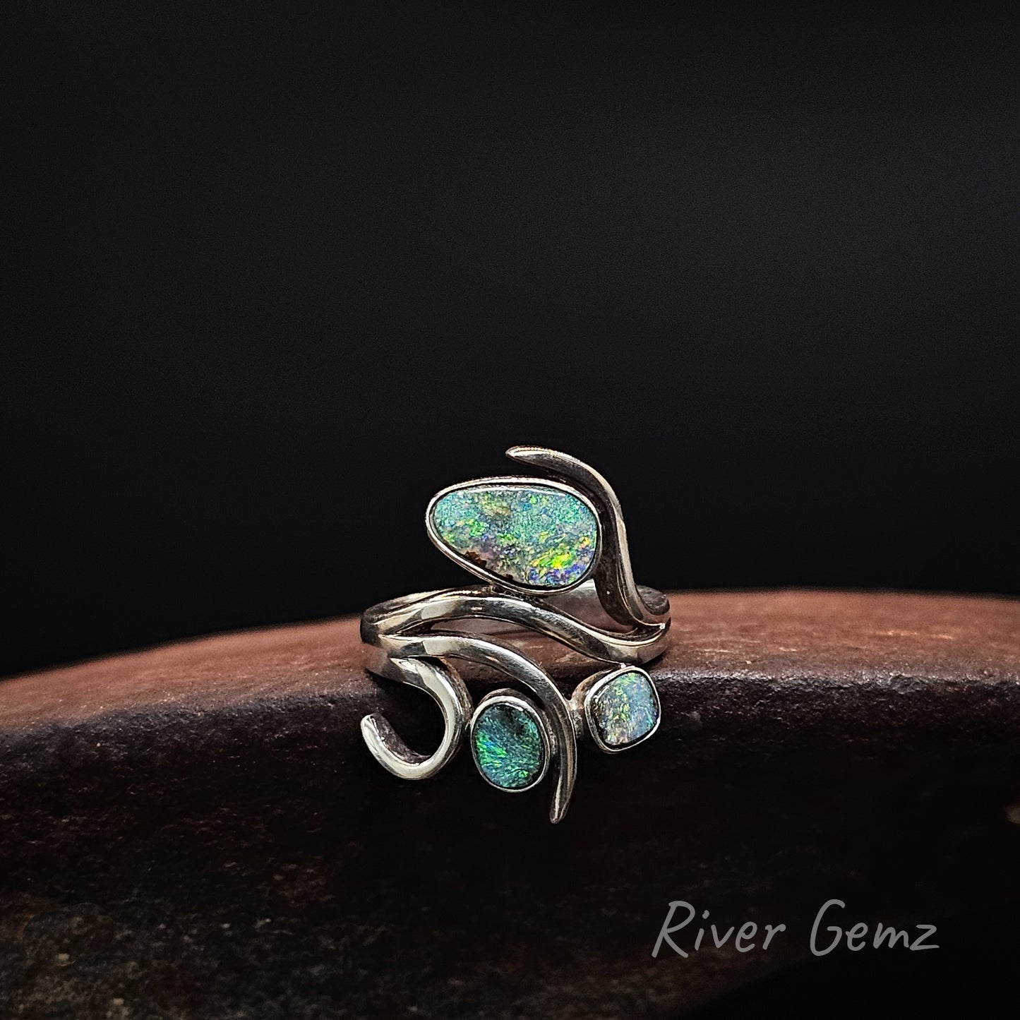 A silver wrap ring featuring three iridescent opal gemstones in organic shapes, displayed on a dark wooden surface against a black background. The brand name 'River Gemz' is visible in the bottom right corner.