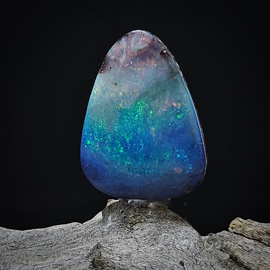 Rounded triangle shaped boulder opal with pinks at top morphing into green and then blue. Opal is upright on a light-grey grained piece of wood and a black backdrop.