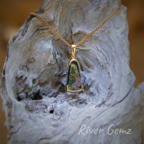 Rounded-triangle shaped multi-coloured pendant partial besel set in 14K gold. Necklace is photographed hanging from a light-grey weathered piece of driftwood.